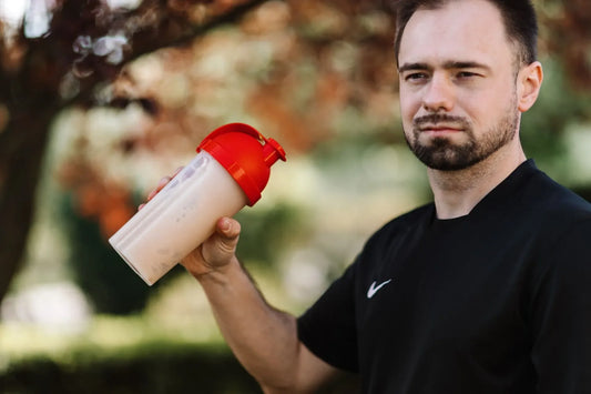 Adult man wearing a black shirt holds a protein shake in an outdoor setting with a blurred nature background